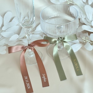 May include: A tall clear glass flute and a wide clear ribbed coupe glass, each adorned with a satin ribbon. The copper ribbon on the flute features the white text Sophie and 28.11.2026. The sage green ribbon on the coupe features the gold text KAROLA.