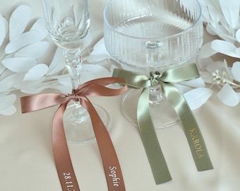 Puede incluir: Una flauta de cristal transparente y una copa ancha de cristal estriado, cada una adornada con una cinta de satén. La cinta de color cobre en la flauta lleva el texto blanco Sophie y 28.11.2026. La cinta verde salvia en la copa lleva el texto dorado KAROLA.