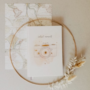 Könnte beinhalten: Ein weißes Notizbuch mit den Worten "collect moments" und einer goldenen Kamera-Illustration. Das Notizbuch ist von einem goldenen Reifen, einer Karte und Trockenblumen umgeben. Die Karte hat ein Vintage-Design.