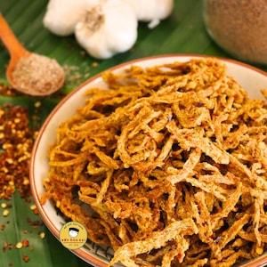 May include: A bowl of golden-brown, shredded food, possibly a snack or appetizer, sits on a green leaf. Garlic cloves, a wooden spoon with seasoning, and red pepper flakes are also visible. The food appears crispy and textured, with a savory appearance.