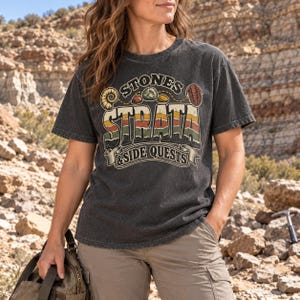以下が含まれることがあります： 「STONES STRATA & SIDE QUESTS」というテキストがプリントされた、チャコールグレーの半袖Tシャツ。デザインには、地層をイメージしたレイヤードスタイルで、落ち着いたアースカラーの化石や岩石のイラストが描かれています。