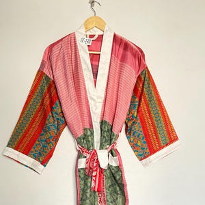 Pode incluir: Uma bata de seda rosa e verde com colarinho branco e padrão floral. A bata tem uma faixa vermelha e branca.