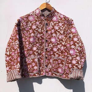 Pode incluir: Uma jaqueta acolchoada marrom com estampa floral em tons de rosa e branco. A jaqueta tem gola mandarim, fecho de botão e dois bolsos de remendo. Os punhos e a bainha são guarnecidos com tecido listrado branco e marrom.