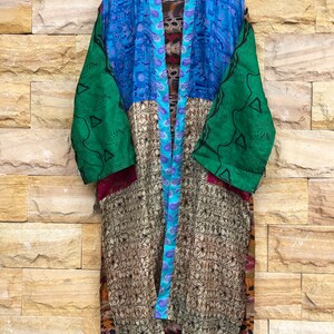 Könnte beinhalten: Ein farbenfroher Patchwork-Kimono-Mantel mit einer Vielzahl von Mustern und Farben, darunter Blau, Grün und Gold. Der Mantel hängt an einem Holzbügel vor einer Backsteinmauer. Das Etikett lautet "AF-76".