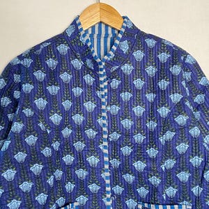 May include: A quilted jacket in a vibrant blue hue, adorned with a repeating floral pattern in light blue and green. The jacket features a mandarin collar, a button-up front, and two pockets. The lining has a blue and white striped pattern.