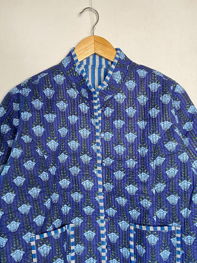 May include: A quilted jacket in a vibrant blue hue, adorned with a repeating floral pattern in light blue and green. The jacket features a mandarin collar, a button-up front, and two pockets. The lining has a blue and white striped pattern.