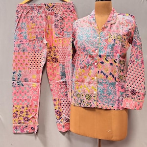 May include: Pink floral pyjama set. The set includes a long-sleeved button-down shirt and matching trousers. The fabric features a patchwork design with various floral and geometric patterns in shades of pink, blue, yellow, and purple.