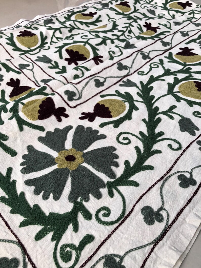 May include: White cotton fabric with a green, brown, and white embroidered floral pattern. The design features stylized flowers and leaves.