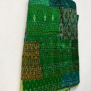 May include: A green kantha quilt with a patchwork design. The quilt is made of different shades of green fabric and is hand-stitched with a running stitch. The quilt is folded and displayed on a white background.