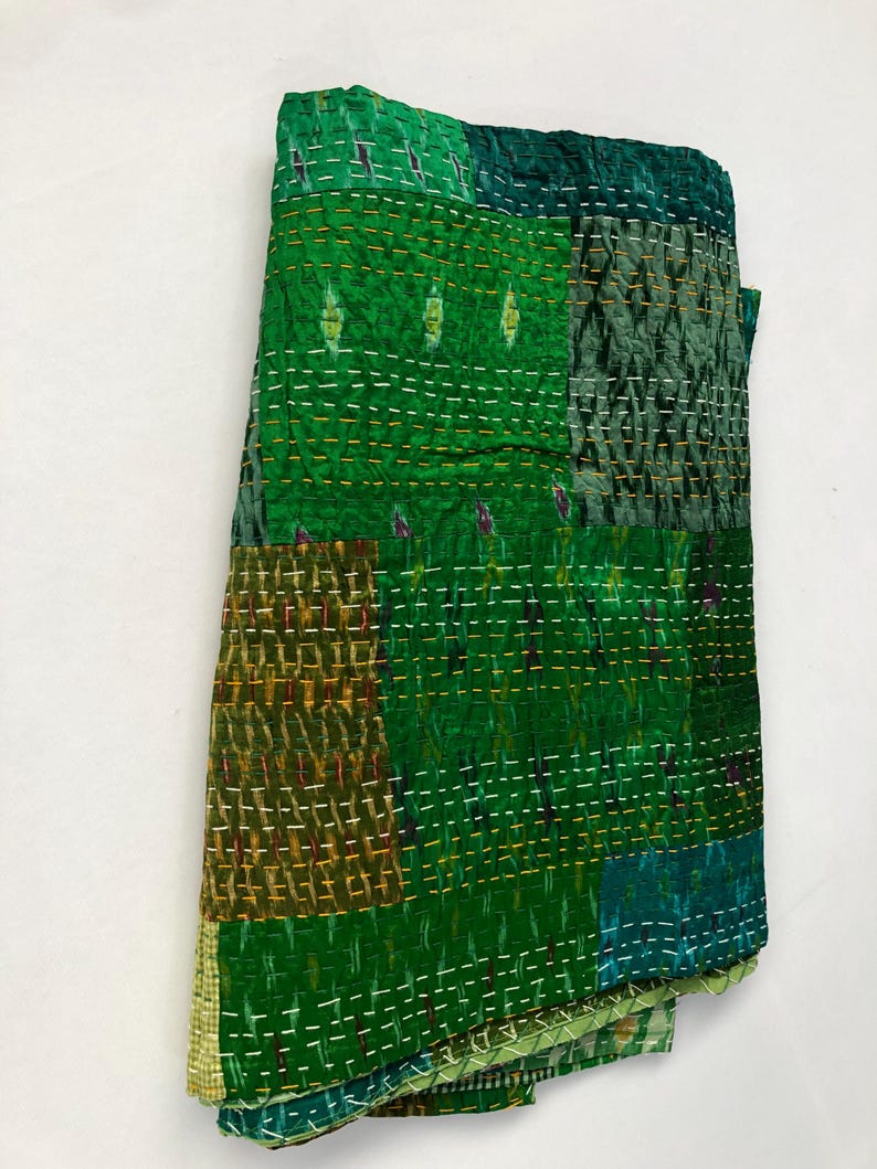 May include: A green kantha quilt with a patchwork design. The quilt is made of different shades of green fabric and is hand-stitched with a running stitch. The quilt is folded and displayed on a white background.
