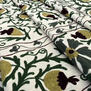 May include: White fabric with green, burgundy, and yellow embroidered floral designs. The fabric is folded in layers, showing the intricate details of the embroidery.