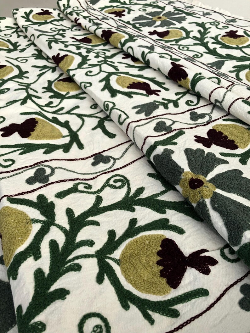 May include: White fabric with green, burgundy, and yellow embroidered floral designs. The fabric is folded in layers, showing the intricate details of the embroidery.