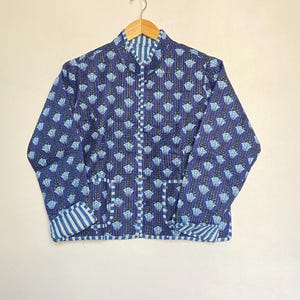 May include: A blue quilted jacket with a floral pattern and striped accents. The jacket features a stand-up collar, two pockets, and a button-up front. The jacket is hanging on a wooden hanger.
