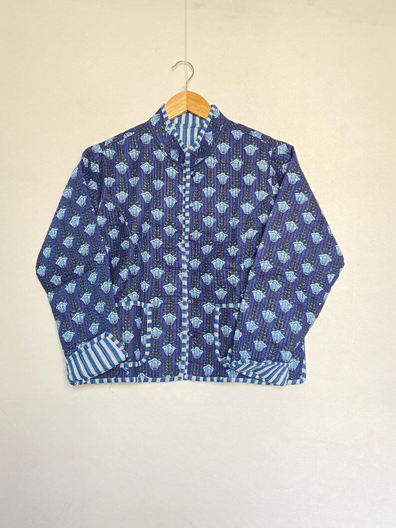 May include: A blue quilted jacket with a floral pattern and striped accents. The jacket features a stand-up collar, two pockets, and a button-up front. The jacket is hanging on a wooden hanger.