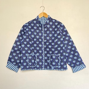 May include: A blue quilted jacket with a floral pattern. The jacket has a mandarin collar, two front pockets, and striped cuffs. The jacket is hanging on a wooden hanger.