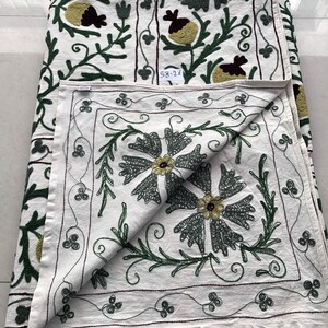 May include: White cotton tablecloth with green and brown embroidery of floral designs. The tablecloth has a square shape and is folded in half. The embroidery is detailed and intricate, with a variety of different flowers and leaves. The tablecloth is perfect for adding a touch of elegance to any table setting.