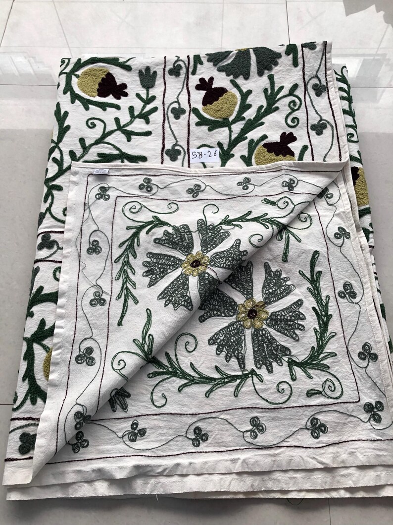 May include: White cotton tablecloth with green and brown embroidery of floral designs. The tablecloth has a square shape and is folded in half. The embroidery is detailed and intricate, with a variety of different flowers and leaves. The tablecloth is perfect for adding a touch of elegance to any table setting.
