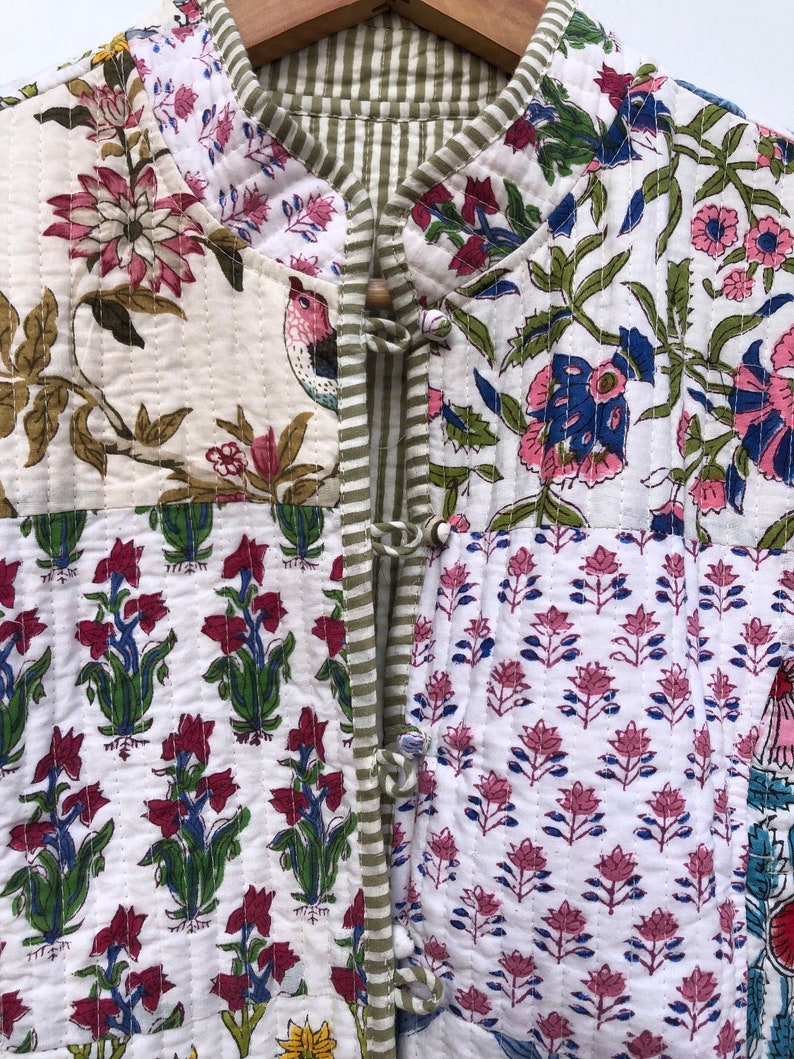 Op de afbeelding: Een wit gewatteerd jack met een bloemenprint. Het jack heeft een mandarijnskraag en een knoopsluiting. De stof is een patchwork van verschillende bloemenpatronen in tinten roze, blauw, groen en rood.
