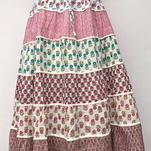 Pode incluir: Uma saia de várias camadas com estampa floral em tons de rosa, branco, verde e marrom. A saia tem cintura com cordão e uma bainha branca.