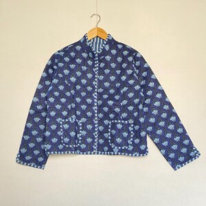 May include: A blue quilted jacket with a floral pattern and two front pockets. The jacket has a stand-up collar and is hanging on a wooden hanger. The jacket's trim and interior lining feature a striped pattern.