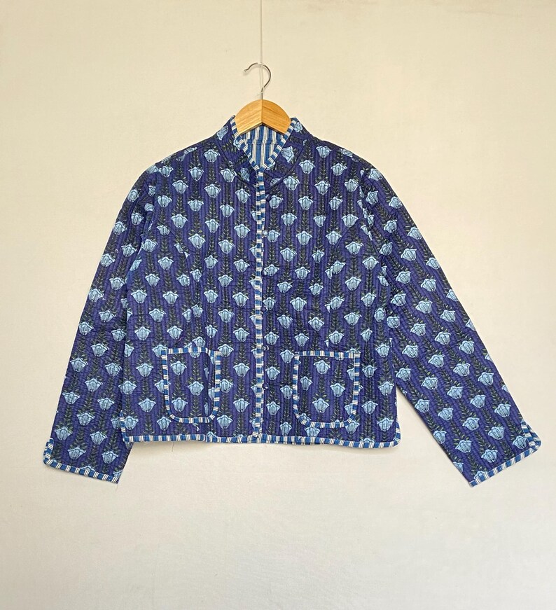 May include: A blue quilted jacket with a floral pattern and two front pockets. The jacket has a stand-up collar and is hanging on a wooden hanger. The jacket's trim and interior lining feature a striped pattern.