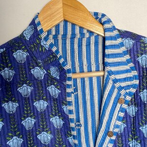 May include: A reversible quilted jacket with a floral pattern on a navy blue background. The jacket features a striped interior in shades of blue and white. It has wooden buttons and is hanging on a wooden hanger.