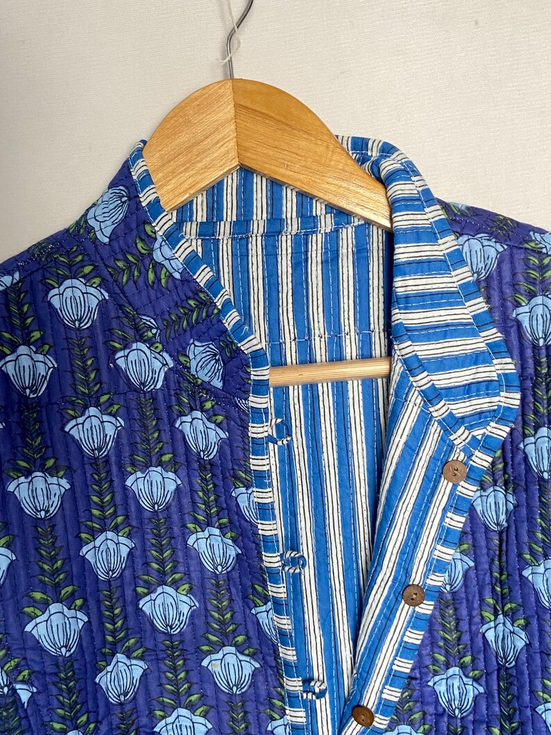 May include: A reversible quilted jacket with a floral pattern on a navy blue background. The jacket features a striped interior in shades of blue and white. It has wooden buttons and is hanging on a wooden hanger.