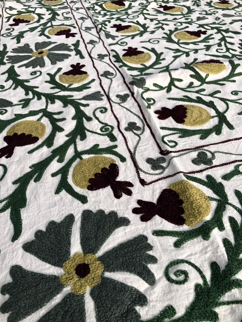 May include: White fabric with intricate embroidery in shades of green, yellow, and burgundy. The design features stylized flowers and leaves.