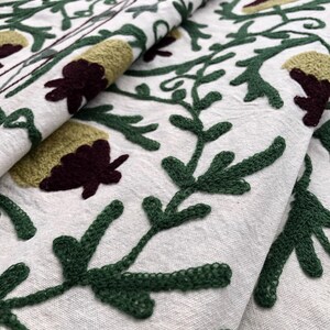 May include: Close-up of a white fabric with green and burgundy embroidery. The embroidery features a floral pattern with leaves and flowers.