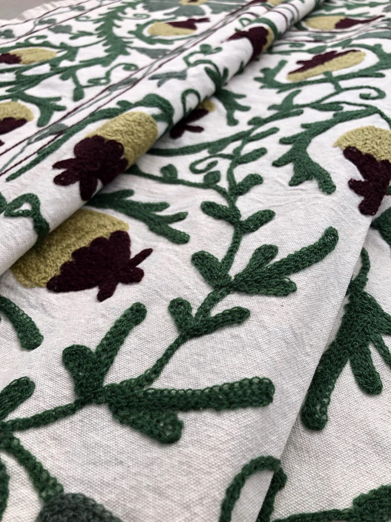 May include: Close-up of a white fabric with green and burgundy embroidery. The embroidery features a floral pattern with leaves and flowers.