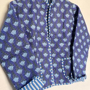 May include: A quilted jacket in shades of blue with a floral pattern. The jacket features a mandarin collar, a button-up front, and striped accents on the cuffs and inner lining. The jacket has two pockets and is approximately 28 inches long.