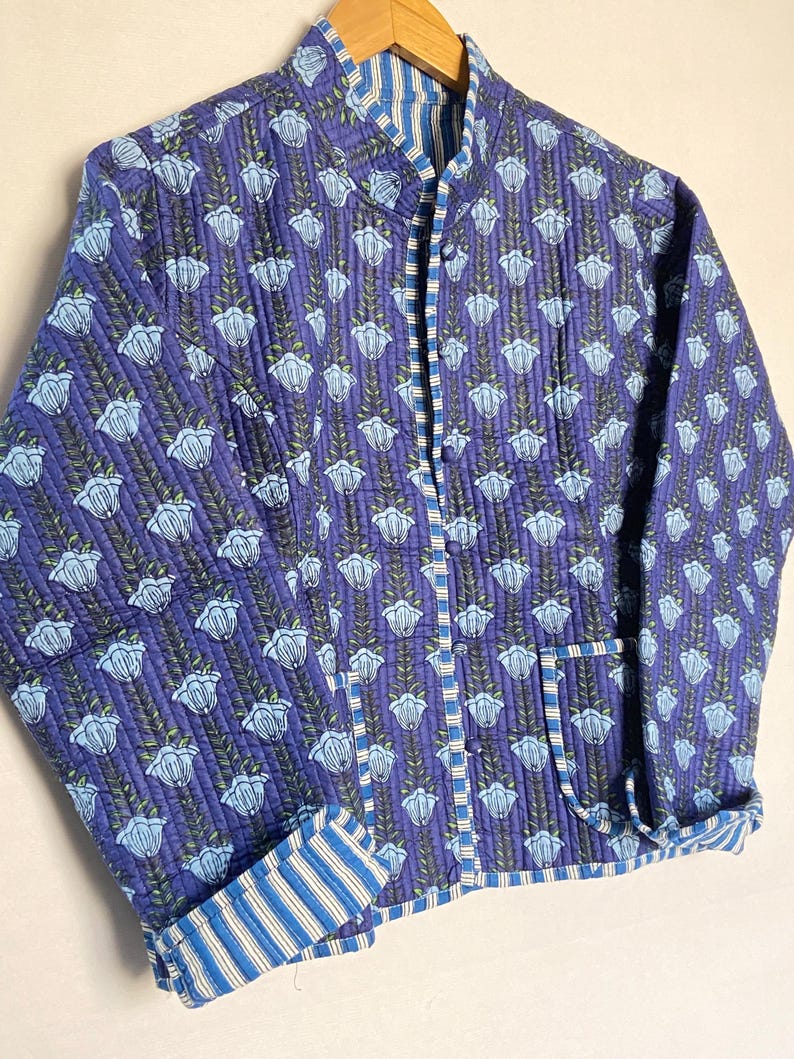 May include: A quilted jacket in shades of blue with a floral pattern. The jacket features a mandarin collar, a button-up front, and striped accents on the cuffs and inner lining. The jacket has two pockets and is approximately 28 inches long.
