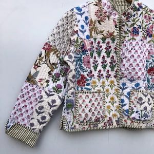 Op de afbeelding: Een wit gewatteerd jack met een bloemenpatchwork-ontwerp. Het jack heeft een mandarijnsluiting, een ritssluiting en twee klepzakken. De stof is een mix van katoen en zijde.
