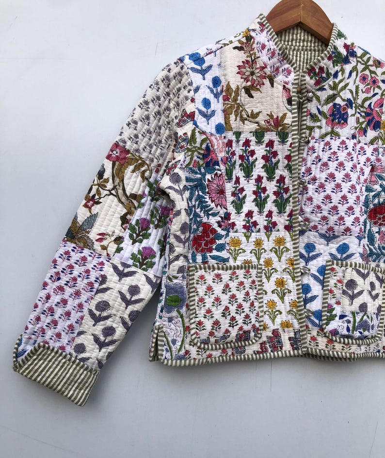 Op de afbeelding: Een wit gewatteerd jack met een bloemenpatchwork-ontwerp. Het jack heeft een mandarijnsluiting, een ritssluiting en twee klepzakken. De stof is een mix van katoen en zijde.