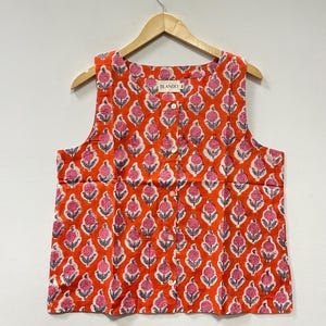 Könnte beinhalten: Ärmelloses orangefarbenes Oberteil mit einem sich wiederholenden Blumenmuster in Rosa, Weiß und Grau. Das Oberteil hat eine Knopfleiste vorne und hängt an einem Holzbügel. Der Markenname "BLANDO" ist auf dem Oberteil aufgedruckt.
