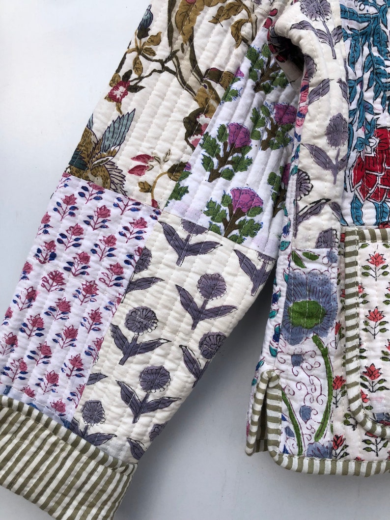 Op de afbeelding: Een wit gewatteerd jack met een patchwork-ontwerp met bloemenpatronen in tinten roze, blauw, groen en bruin. Het jack heeft een groen-wit gestreepte afwerking.