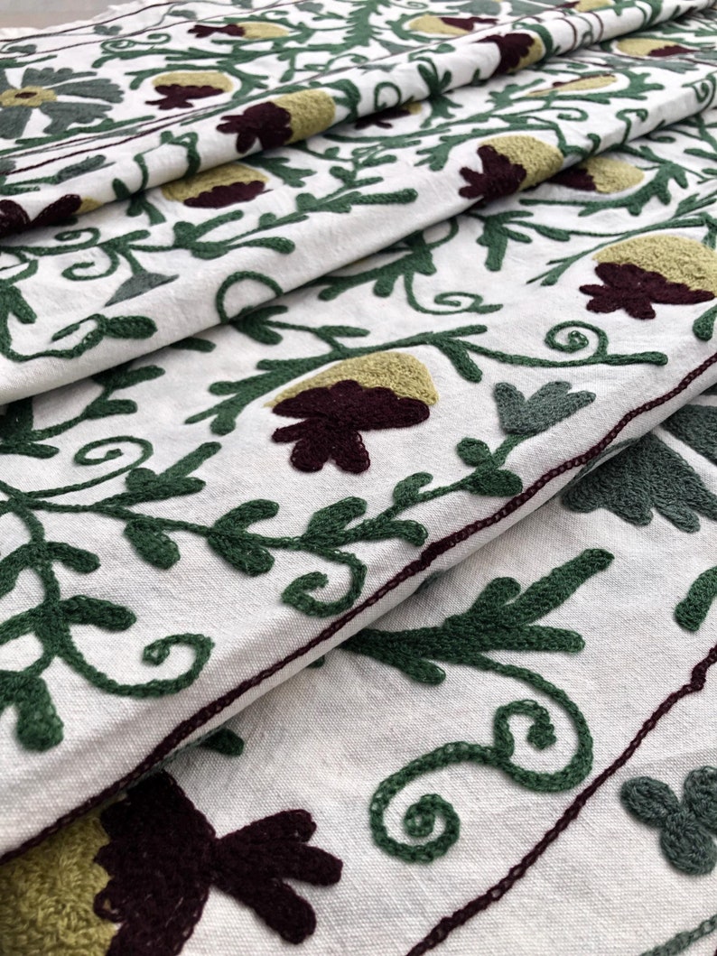 May include: White fabric with green and burgundy embroidery of floral designs. The fabric is folded in layers, showing the intricate details of the embroidery.