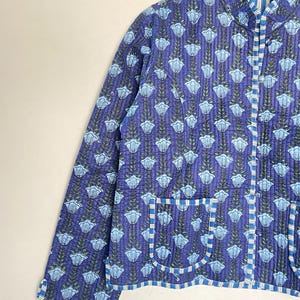 May include: A quilted jacket in shades of blue with a floral pattern. The jacket features a mandarin collar, long sleeves, and two front pockets. The edges are trimmed with a striped fabric. The jacket is approximately 25 inches long.