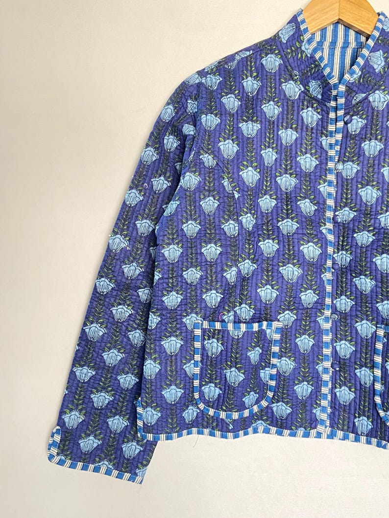 May include: A quilted jacket in shades of blue with a floral pattern. The jacket features a mandarin collar, long sleeves, and two front pockets. The edges are trimmed with a striped fabric. The jacket is approximately 25 inches long.