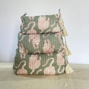 May include: Three stacked, rectangular cosmetic bags in varying sizes. The bags are sage green with a repeating pattern of white tigers. Each bag has a zipper closure and a small tassel. The bags are stacked on top of each other.