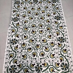 May include: White cotton throw blanket with a hand-stitched design of green leaves and burgundy pomegranates. The design is repeated in a border around the blanket.