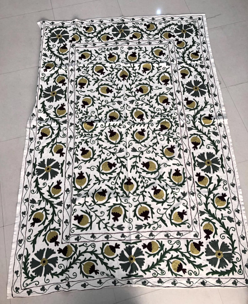 May include: White cotton throw blanket with a hand-stitched design of green leaves and burgundy pomegranates. The design is repeated in a border around the blanket.