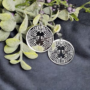 May include: A pair of silver earrings with a black and white circuit board design. The earrings are round and have a silver hook.