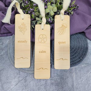 May include: Three wooden bookmarks with cream-colored ribbons. Each bookmark features a different design: a coral, a sand dollar, and a jellyfish. The words "steady", "calm", and "quiet" are engraved below the designs, with a wave graphic at the bottom.