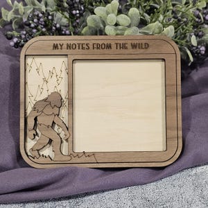 May include: Wooden frame with the text "MY NOTES FROM THE WILD". The frame features a Bigfoot silhouette and forest scene on the left, with a blank space on the right. The frame is made of light brown wood.