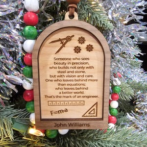 May include: Wooden Christmas ornament with an arched top, featuring engraved text and engineering symbols. The text reads "Someone who sees beauty in precision...That's the mark of an engineer." Includes the name "John Williams".