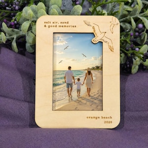 May include: A rectangular light-colored wooden picture frame featuring an engraved seagull design in the top right corner. The frame includes the text "salt air, sand & good memories" at the top and "orange beach 2026" at the bottom right.