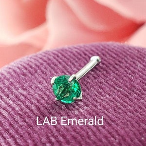 May include: A silver nose ring with a green lab-created emerald gemstone. The ring is on a green velvet surface. LAB Emerald.