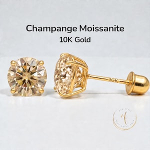 May include: A pair of champagne moissanite stud earrings set in 10K gold. The round-cut stones are held by four-prong settings, with the post and screw-back closure clearly marked with 10K. The text Champange Moissanite 10K Gold is visible.