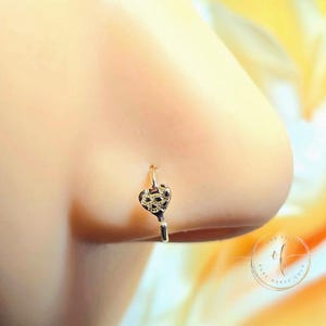 May include: A gold-colored nose ring with a heart-shaped charm. The charm has a textured design and hangs from a small hoop that fits around the nostril. The background is blurred, with a hint of yellow and white.